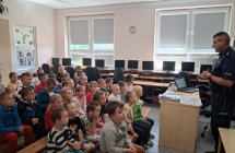Uczniowie klas 0-3 siedzacy na lawkach podczas spotkania z policjantem